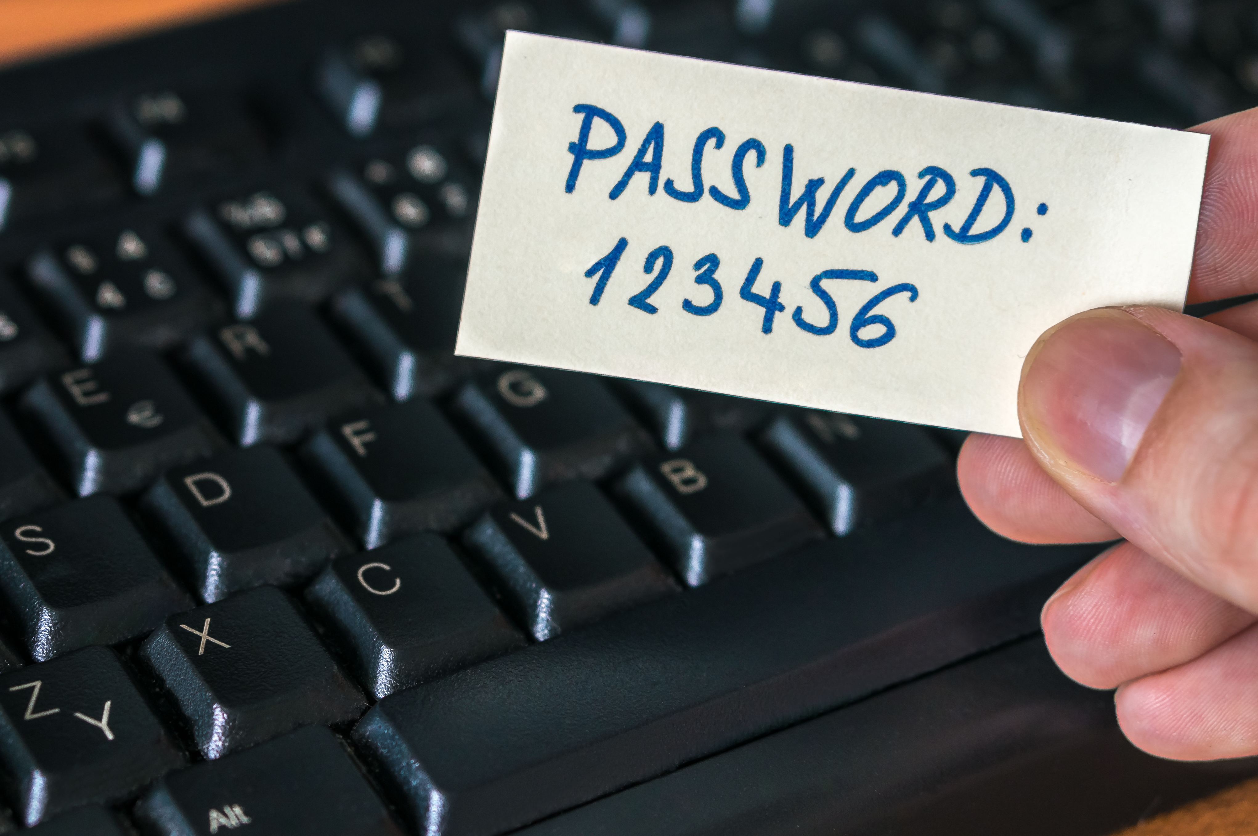 The image shows a person’s hand hovering over a computer keyboard. They are holding a piece of paper with the password 123456 written on it.