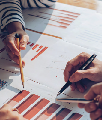 Four hands holding pens/pencils and examining graphs and spreadsheets